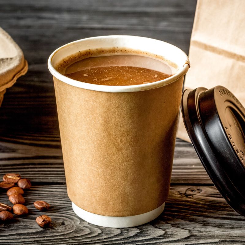 Brown paper cup with coffee on a wooden surface with coffee beans and a brown bag.