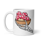 White mug with a skeleton hand holding a bowl of red and white polka dot mushrooms design.