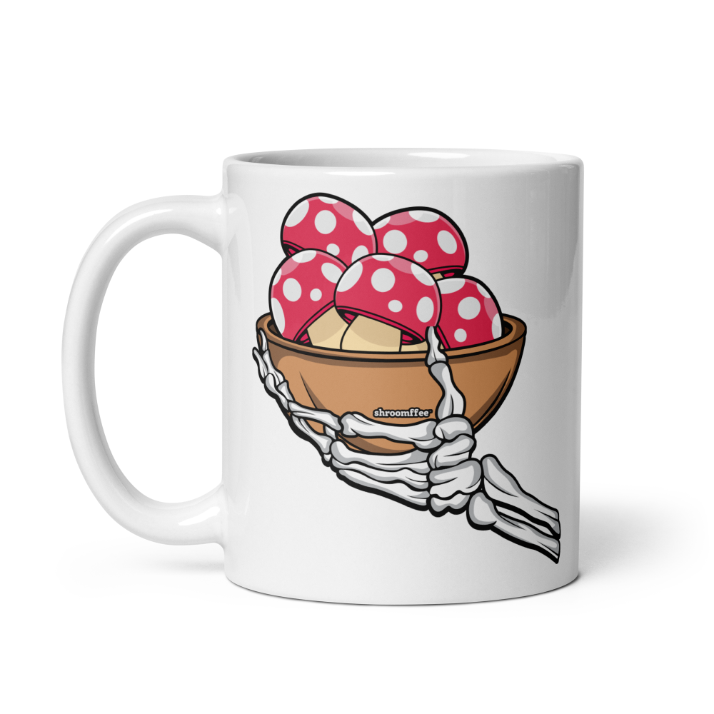 White mug with a skeleton hand holding a bowl of red and white polka dot mushrooms design.