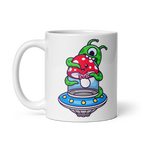 A white ceramic mug with a colorful illustration of an alien inside a flying saucer about to eat a red and green mushroom.